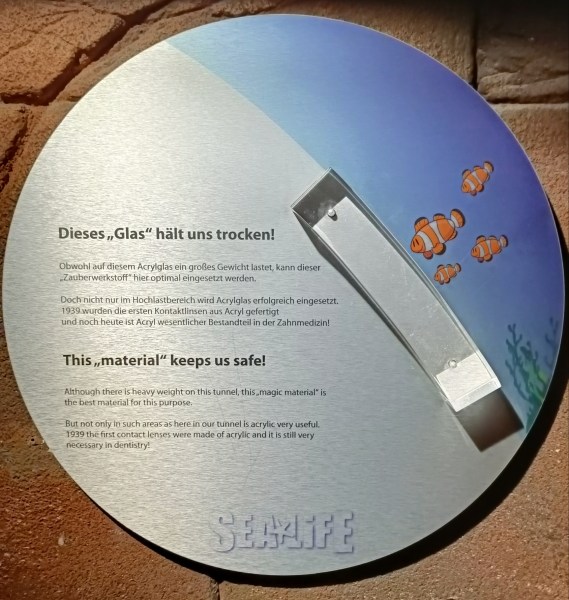 Info-Tafel des Sealife Hannover zum Acrylglas der Aquarien, auf der seltsamerweise die Ausdrücke »Glas« und in der englischen Version »material« in Anführungszeichen gesetzt sind.