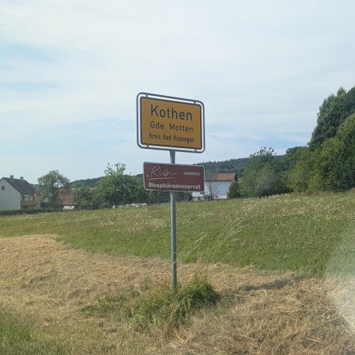 Bild des Ortseingangsschilds des Ortes Kothen, Gemeinde Motten, Kreis Bad Kissingen. Im Hintergrund ein paar Häuser